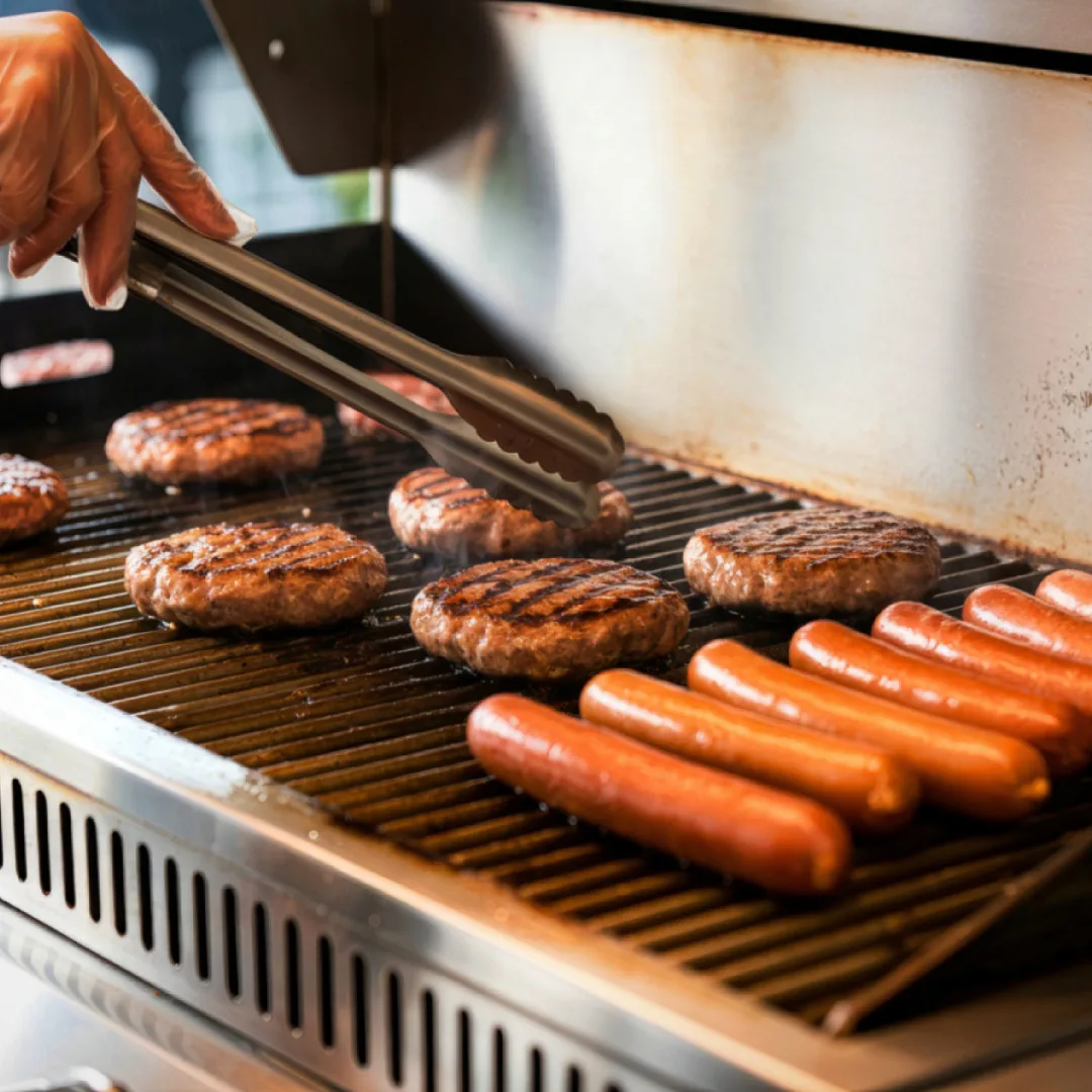Chef grilling multiple beef patties and hot dogs on commercial grill grates, demonstrating high-volume foodservice preparation and wholesale bulk quan