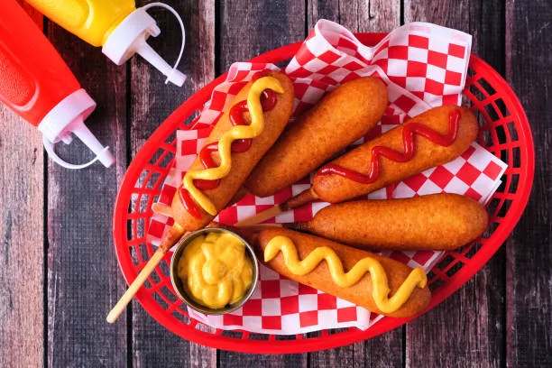 Golden breaded corn dogs with mustard drizzle in red checkered foodservice basket with condiment packets for bulk distribution.